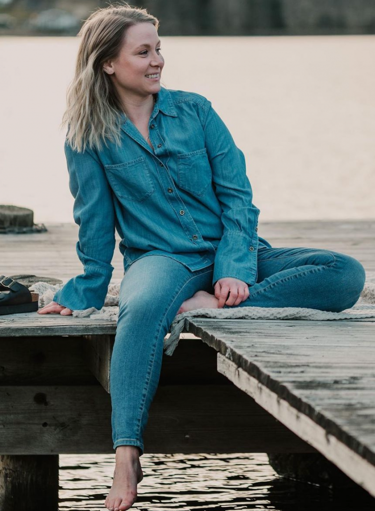 Influencern Theodora Larsson sitter avslappnat på en brygga vid en sjö med stilla vatten runt omkring.