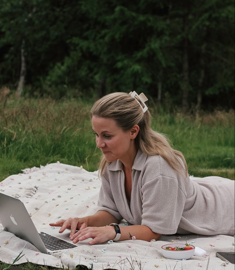 Influencer Theodora Larsson ligger på en filt i gräset och skriver på en bärbar dator.