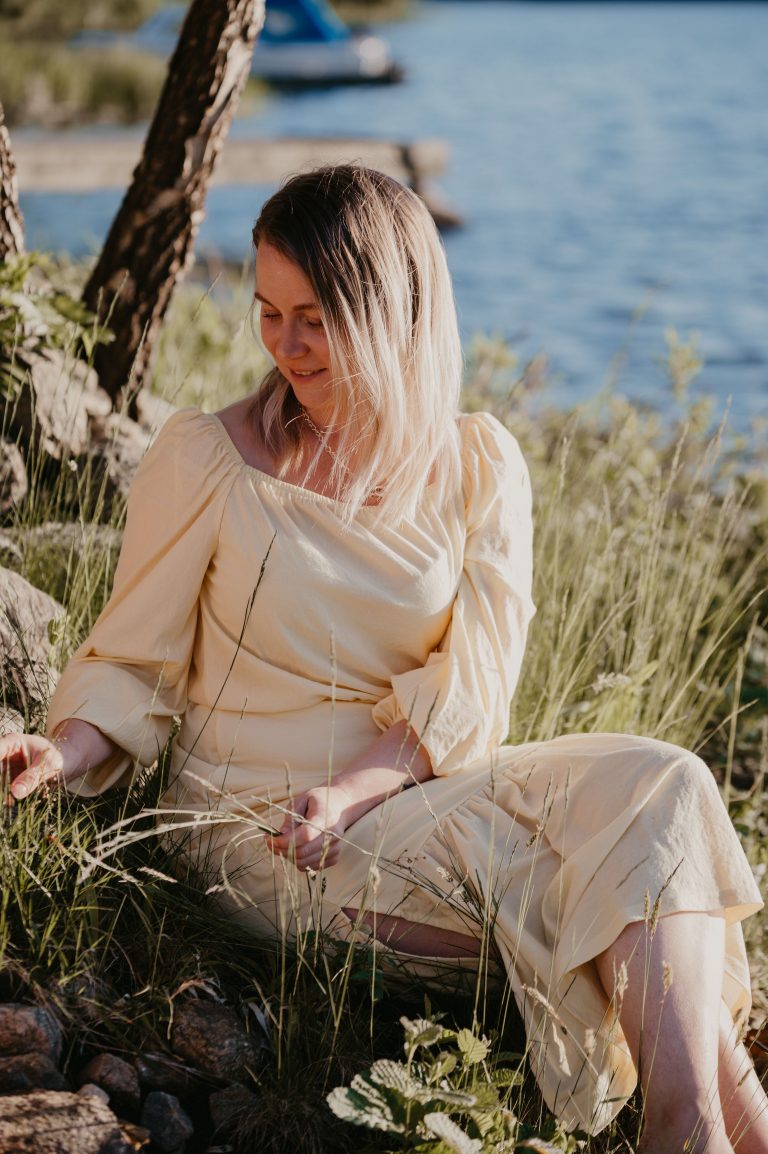 Influencer Theodora Larsson sitter i gräset en solig dag med vatten i bakgrunden.