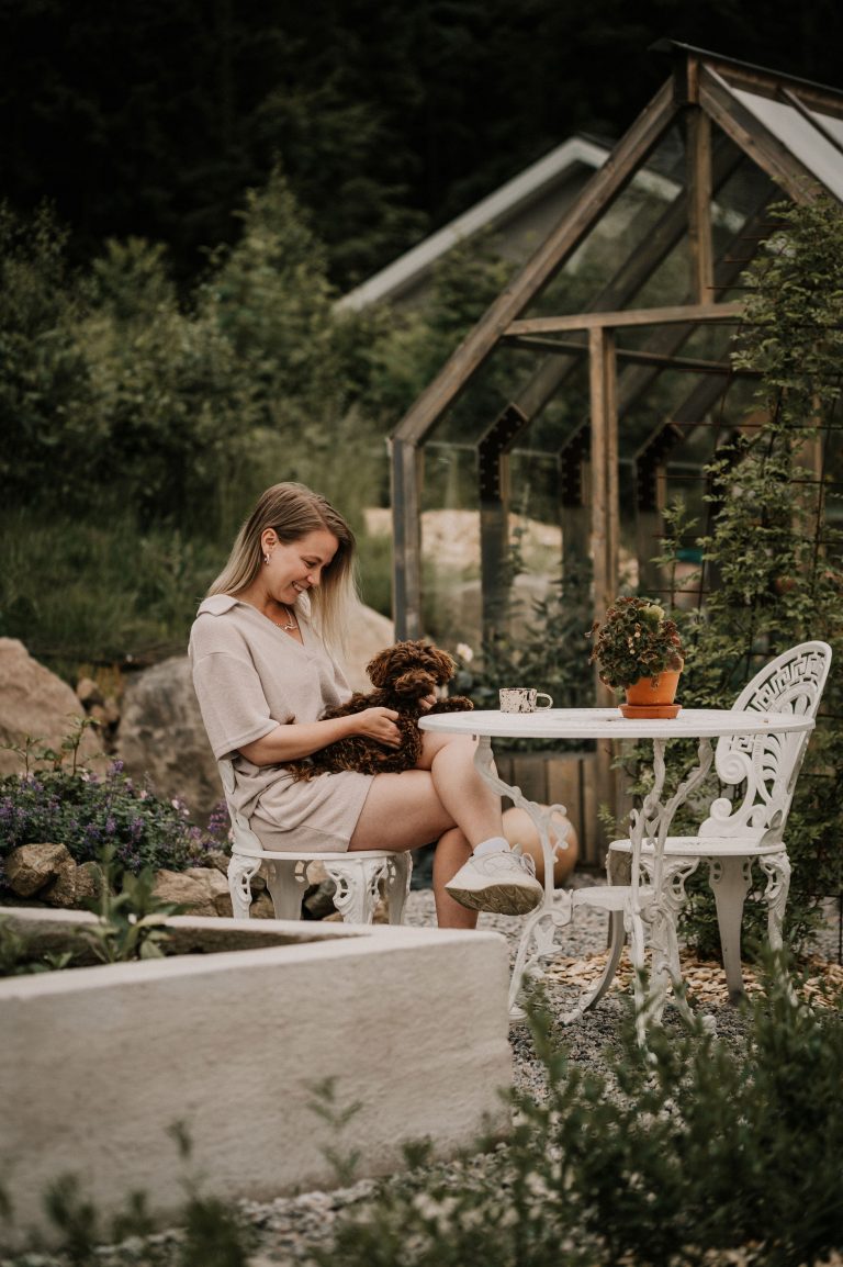 Influencer Theodora Larsson sitter med en brun hund i knät framför ett växthus.