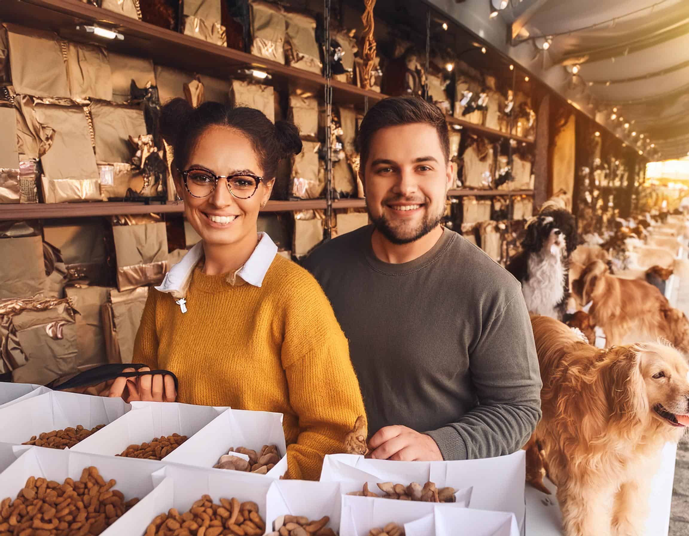 Två människor står i Pawsome Treats monter under en hundmässa. Runt dem finns förpacknignar med hundgodis och bredvid dem står hundar.